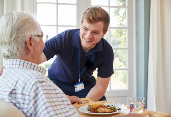 Caregiver smiling at a male resident before he digs into his food.